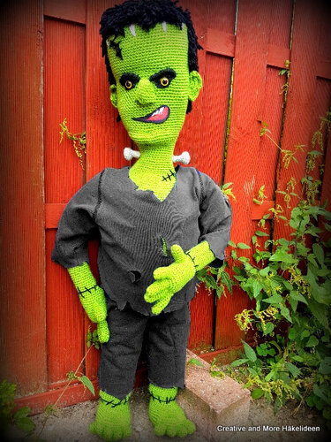 Handgehäkelte grüne Frankenstein‑ähnliche Puppe mit schwarzem Haar und zerrissener Kleidung vor rotem Holzzaun