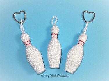 Drei gehäkelte weiße Kegel‑Schlüsselanhänger mit rot‑weißem Zierfaden und zwei herzförmigen Ringen auf hellblauem Hintergrund.
