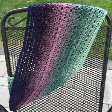 Gehäkelter Loop‑Schal mit Farbverlauf von Dunkelblau bis Türkisgrün über einer Metallstuhllehne im Freien.