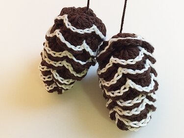 Zwei gehäkelte Tannenzapfen zum Aufhängen, braun mit weißen Schuppenkanten auf hellem Hintergrund.