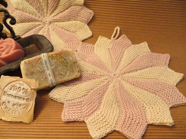 Zwei gestrickte sternförmige Topflappen in Rosa und Creme neben Seifen auf geriffeltem Untergrund.