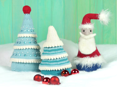 Knitted blue-and-white tiered Christmas trees and a small Santa figure with red hat and white beard, with red baubles on a snowy surface.