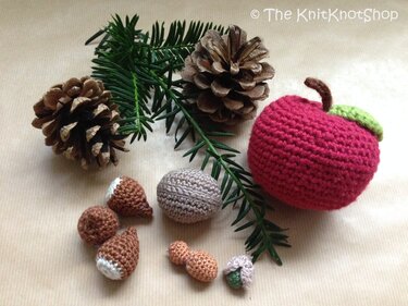 Gehäkelter roter Apfel neben gehäkelten Eicheln und kleinen Kugeln mit Tannenzapfen und Tannenzweig