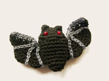 Gehäkelte schwarze Fledermaus-Amigurumi mit roten Perlenaugen und grau akzentuierten Flügeln.