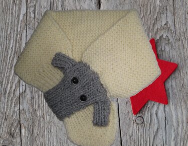 Cremefarbener gestrickter Babyschal mit grauer Tierapplikation und schwarzen Knopfaugen auf Holzuntergrund