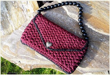 Weinrote gehäkelte Clutch mit schwarzem Perlenhenkel und herzförmigem Knopf auf einer Steinoberfläche