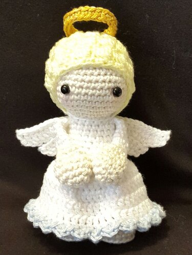 Weißer gehäkelter Amigurumi-Engel mit gelbem Haar, goldfarbenem Heiligenschein und gefalteten Händen vor dunklem Hintergrund.