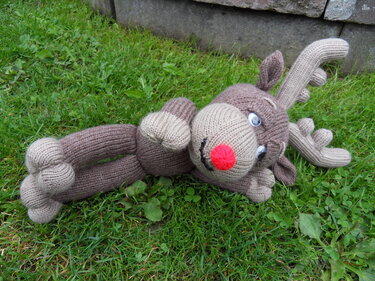Gestricktes braunes Rentier-Amigurumi mit roter Nase liegt auf grünem Gras