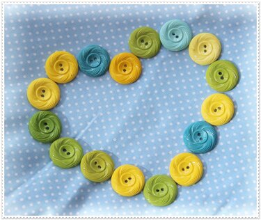 Mehrere gelbe, grüne und türkise Knöpfe mit zwei Löchern zu einer Herzform auf hellblauem Tupfenstoff