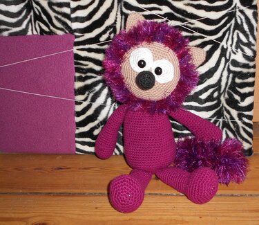 Gehäkelter lila Amigurumi-Löwe mit flauschiger violetter Mähne und buschiger Schwanzspitze vor zebraähnlichem Hintergrund