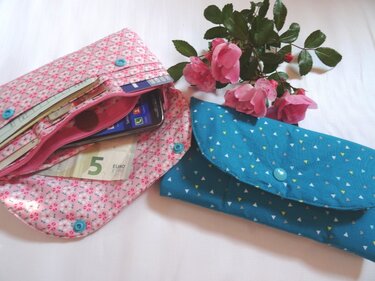 Zwei Stofftaschen: offene rosa Geldbörse mit Karten und geschlossenes türkisfarbenes Etui; Rosen daneben