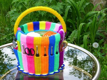 Striped crocheted basket with a yellow handle and external pockets holding scissors and crochet hooks, sitting on a reflective outdoor table amidst green foliage.