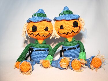 Zwei gehäkelte Kürbispuppen mit blauen Hüten, grünen Ärmeln und Garnhaaren sitzen nebeneinander