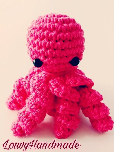 Vorderansicht eines kleinen pinken gehäkelten Amigurumi‑Oktopus mit schwarzen Augen auf hellem Hintergrund.