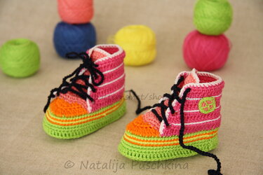 Pair of crocheted baby high-top shoes in pink, orange, green and white with black laces, with yarn balls behind them.