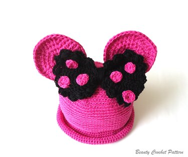 Pink crocheted baby hat with round ears and a black bow with pink polka dots