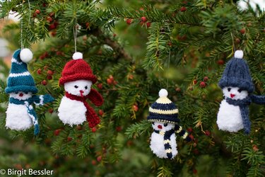 Vier Pompon‑Schneemänner mit gestrickten Mützen und Schals an einem immergrünen Zweig mit roten Beeren