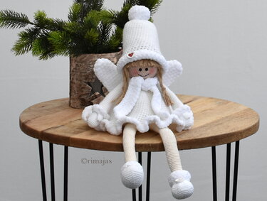 Zarter weißer Amigurumi-Engel mit Bommelmütze und Rüschen sitzt auf Holztisch vor Tannenzweigen