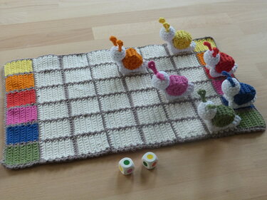 Crocheted board game with six colorful snail figures, grid squares, and two dice on a wooden floor.