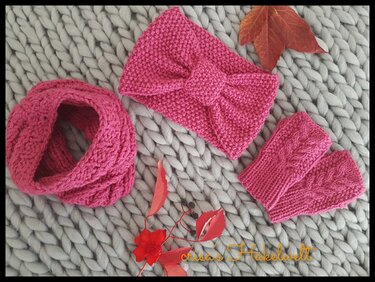 Pinkes Strickset (Loop‑Schal, geknotetes Stirnband, fingerlose Pulswärmer) auf hellgrauer, grober Strickdecke mit roten Herbstblättern.