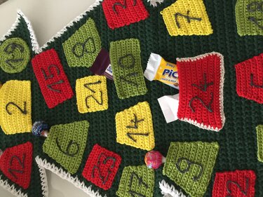 Gehäkelter Adventskalender dunkelgrün mit roten, gelben und grünen Taschen und aufgestickten Zahlen; einige Süßigkeiten sichtbar