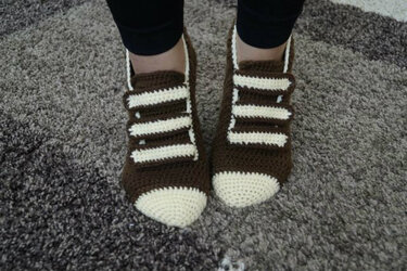 Gehäkelte braun-weiße Hausschuhe mit drei hellen Riemen, getragen auf hellgrauem Teppich