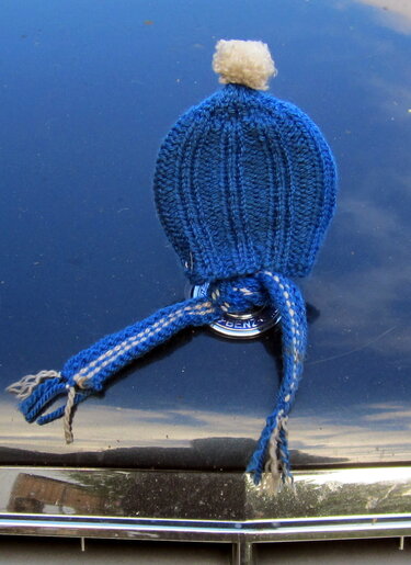 kleine blaue gestrickte mütze mit weißem bommel auf chromfarbenem autoemblem