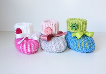 Drei gestrickte Babyschuhe in Pink-Weiß, Hellrosa-Hellgrau und Hellblau-Lindgrün mit Schleifen und Blumenknöpfen auf weißem Hintergrund.