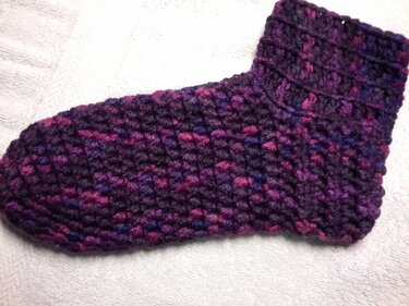 Gehäkelte Socke in melierten Lila- und Pinktönen auf weißem Handtuch