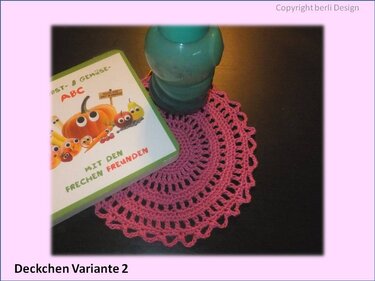Pinkes rundes Häkeldeckchen mit Lochmuster und Wellenrand unter Glas und Buch