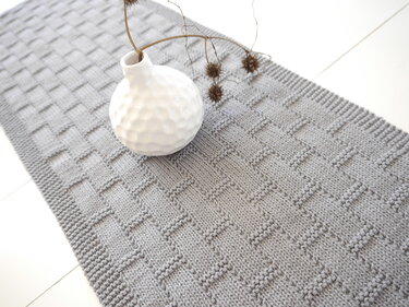 Grau gestrickter Tischläufer mit Korbmuster und weißer Vase mit Trockenblumen