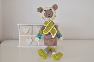 Gehäkelter Amigurumi-Teddybär mit braun-beigem Körper, gestreiften Gliedmaßen und hellgrünem Schal, sitzend vor einer weißen Kommode.
