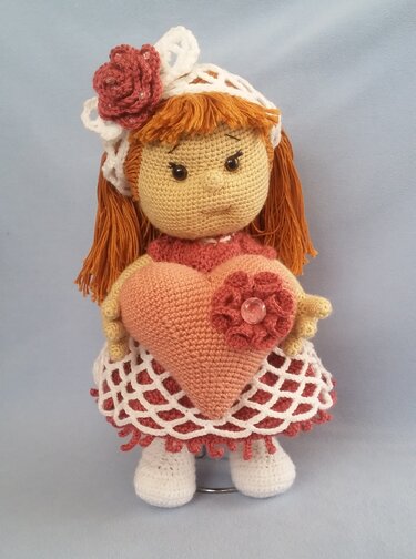Gehäkelte Amigurumi-Puppe mit rostbraunem Haar, Spitzenkleid und gehäkeltem Herz.