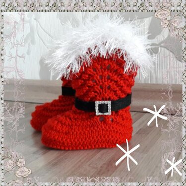 Zwei rote, gestrickte Babyschuhe im Weihnachtsmann-Look mit weißem Plüschrand und schwarzem Gürtel mit Zierschnalle auf Holzuntergrund.