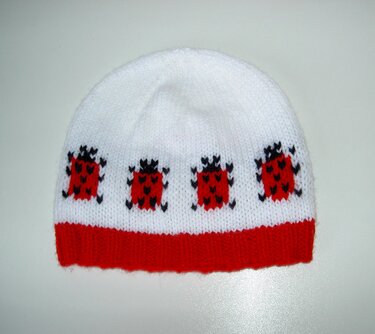 Weiße gestrickte Babymütze mit rotem Rippbündchen und vier roten Käfermotiven.