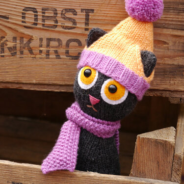 Black amigurumi cat wearing a yellow pompom hat and lavender scarf in a wooden crate