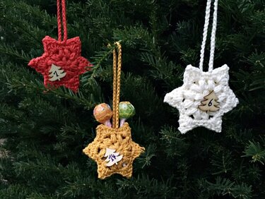 Drei kleine gehäkelte Stern-Taschen mit Schlaufen und nummerierten Holzplättchen, an einem Weihnachtsbaum aufgehängt; eine Tasche enthält Lollis