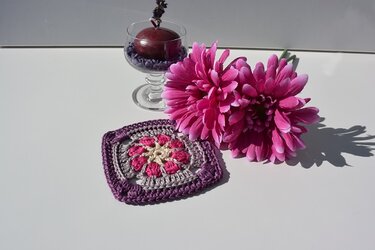 Quadratisches gehäkeltes Granny Square in Lila, Grau, Pink und Creme, daneben pinkfarbene Blumen und eine Kerze in einem Glas.