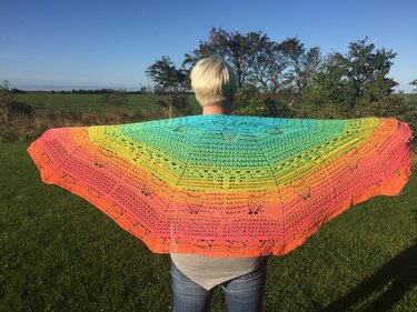 Person von hinten trägt ein weit ausgebreitetes halbrundes Häkel­tuch mit Farbverlauf von Türkis bis Pink vor einer ländlichen Kulisse.