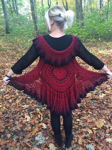 Rückenansicht einer Frau im Wald, trägt eine bordeauxrote bis schwarze gehäkelte Jacke mit rundem Mandala‑Motiv und weitem Saum