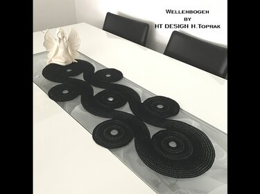 Black crochet table runner of linked circular motifs forming an S-shaped wave on a white dining table with glass inset