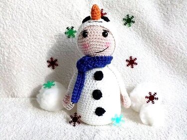 Gehäkelter Amigurumi‑Schneemann mit blauem Schal, Karottennase und schwarzen Knöpfen auf weißer Oberfläche