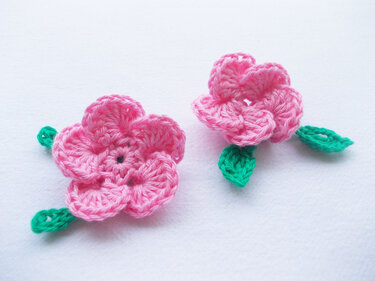 zwei rosa gehäkelte Blumenapplikationen mit grünen Blättern auf weißem Hintergrund