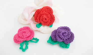 Drei gehäkelte Blumenapplikationen in Rot, Pink und Lila mit grünen Blättern auf hellem Hintergrund.