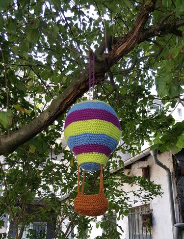 Gehäkelter Heißluftballon in bunten Streifen mit kleinem Korb, an einem Baum hängend