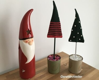 Zwei gehäkelte spitze Weihnachtsbäume auf Stäben in kleinen Bechern neben einer roten Keramikfigur