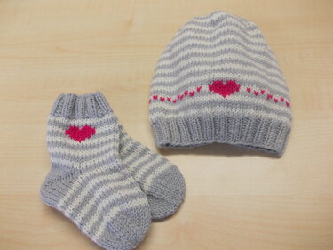 Gestrickte Babymütze und ein Paar Babysocken in grau-weiß gestreift mit pinken Herzmotiven auf hellem Holzuntergrund.
