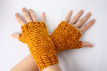 Eine Person trägt senfgelbe, gestrickte fingerlose Handschuhe an beiden Händen.