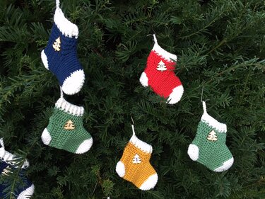 Verschiedene gehäkelte Mini-Socken in Gelb, Grün, Blau und Rot mit weißen Bündchen und nummerierten Holzknöpfen, aufgehängt an Zweigen