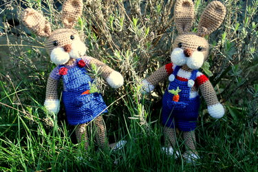 Zwei gehäkelte Amigurumi-Hasen im Gras, einer im blauen Latzkleid, einer in blauer Latzhose